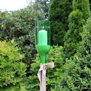 May include: A green glass candle holder made from a repurposed bottle, with the word "FREUDENFEUER" printed on it. A green candle is inside. The holder is tied with a beige ribbon and set against a backdrop of green foliage.
