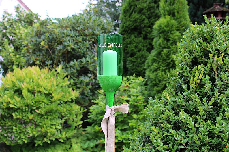 May include: A green glass candle holder made from a repurposed bottle, with the word "FREUDENFEUER" printed on it. A green candle is inside. The holder is tied with a beige ribbon and set against a backdrop of green foliage.