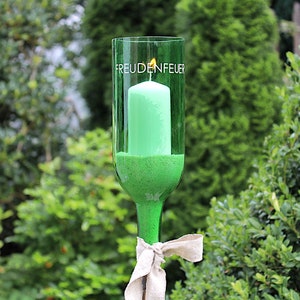 May include: A green glass candle holder with a lit green candle inside. The words "FREUDENFEUER" are printed on the glass. The candle holder is tied with a beige ribbon and set against a backdrop of green foliage.
