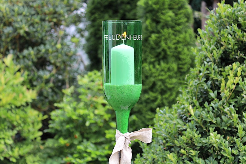 May include: A green glass candle holder with a lit green candle inside. The words "FREUDENFEUER" are printed on the glass. The candle holder is tied with a beige ribbon and set against a backdrop of green foliage.