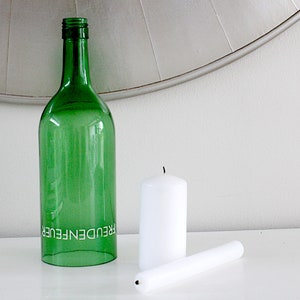 May include: A green glass bottle with the word "FREUDENFEUER" printed on it, next to two white candles. One candle is upright, and the other is lying down. The items are on a white surface.