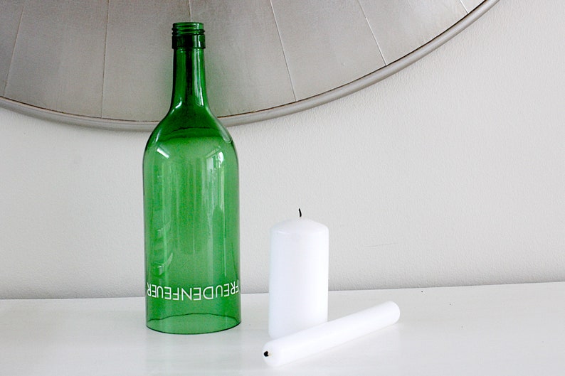 May include: A green glass bottle with the word "FREUDENFEUER" printed on it, next to two white candles. One candle is upright, and the other is lying down. The items are on a white surface.