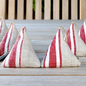 Könnte beinhalten: Sechs Türstopper aus Leinen mit rot-weißen Streifen. Jeder Türstopper ist kegelförmig und hat eine Juteschlaufe. Sie sind auf einer Holzoberfläche angeordnet und bieten eine rustikale, ländliche Ästhetik. Die Höhe beträgt ca. 15 cm.