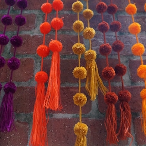 May include: A wall decorated with colorful pom-pom garlands. The garlands are made of fluffy balls of yarn in shades of orange, yellow, purple, and brown. Each garland has a tassel at the end.