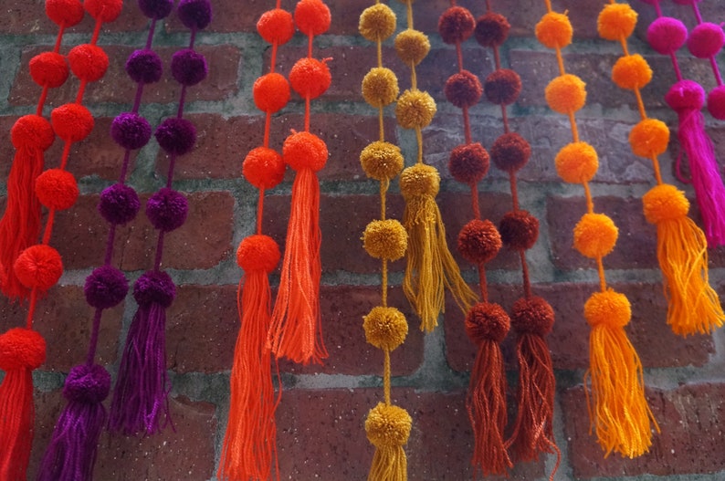 May include: A wall decorated with colorful pom-pom garlands. The garlands are made of fluffy balls of yarn in shades of orange, yellow, purple, and brown. Each garland has a tassel at the end.