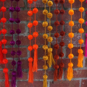 May include: A colorful garland of pom-poms in shades of orange, red, purple, yellow, brown, and pink. The garland is made of yarn and has tassels at the bottom.