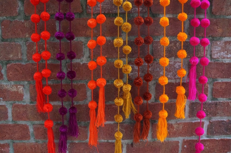 May include: A colorful garland of pom-poms in shades of orange, red, purple, yellow, brown, and pink. The garland is made of yarn and has tassels at the bottom.