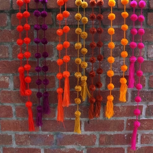 May include: A garland of colorful pom-poms with tassels hanging from a brick wall. The garland is made of orange, yellow, purple, and pink pom-poms.