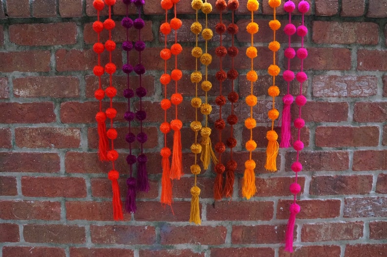 May include: A garland of colorful pom-poms with tassels hanging from a brick wall. The garland is made of orange, yellow, purple, and pink pom-poms.