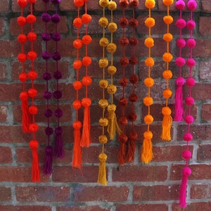 May include: A set of colorful pom-pom garlands with tassels hanging from a brick wall. The garlands are made of yarn in shades of orange, yellow, pink, and purple.