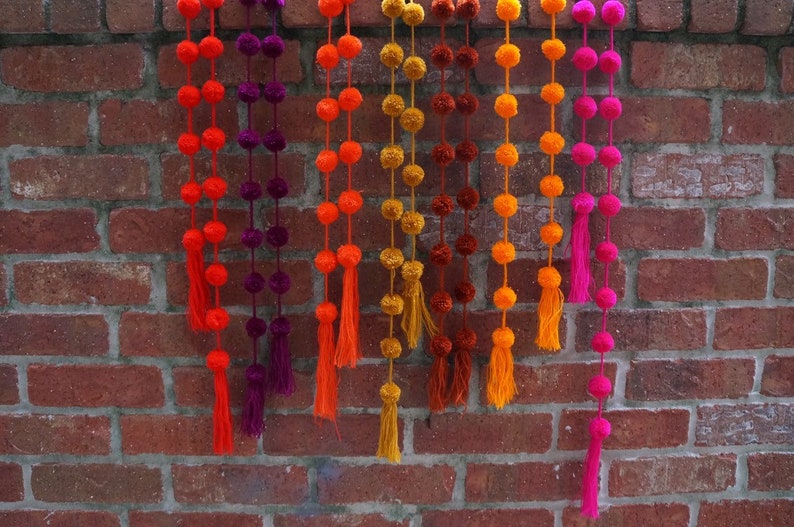 May include: A set of colorful pom-pom garlands with tassels hanging from a brick wall. The garlands are made of yarn in shades of orange, yellow, pink, and purple.