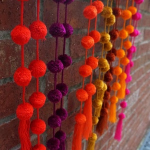 May include: A wall decorated with colorful pom-pom garlands. The garlands are made of fluffy, round pom-poms in shades of orange, red, purple, and gold. The garlands are hung in a staggered pattern, creating a festive and cheerful atmosphere.