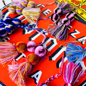 May include: Assortment of colorful tassel ornaments with pom poms. The tassels are made of various yarns in shades of pink, blue, yellow, and purple. The pom poms are made of velvet in shades of brown, pink, and purple.