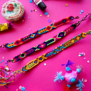 Könnte beinhalten: Drei bunte Freundschaftsarmbänder mit komplizierten gewebten Mustern in Rot-, Gelb-, Blau-, Grün- und Rosatönen. Die Armbänder liegen auf einer leuchtend rosa Oberfläche mit Konfetti, das um sie herum verstreut ist.