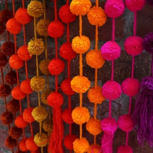 May include: A garland of colorful pom-poms in shades of orange, red, pink, and purple. The pom-poms are strung together on a white string and hang from a brick wall.