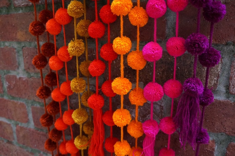 May include: A garland of colorful pom-poms in shades of orange, red, pink, and purple. The pom-poms are strung together on a white string and hang from a brick wall.