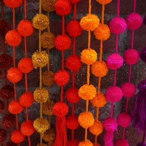 May include: A garland of colorful pom-poms in shades of orange, red, pink, and purple. The pom-poms are strung together on a white string and hang from a brick wall.