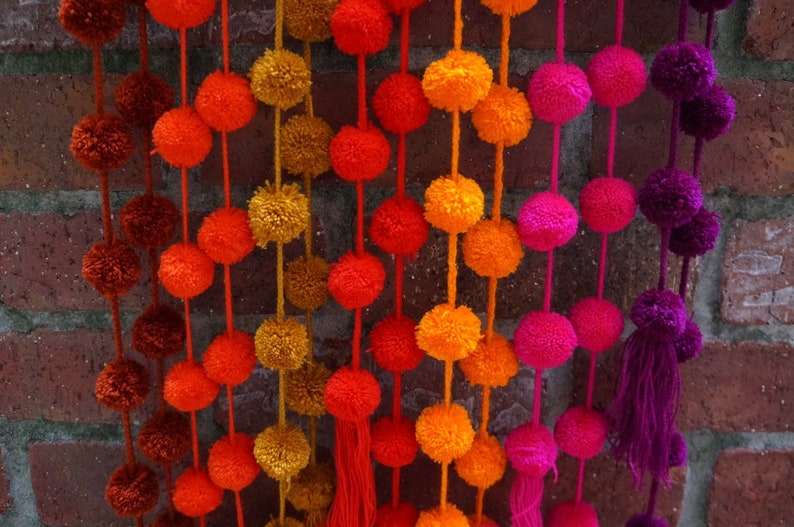 May include: A garland of colorful pom-poms in shades of orange, red, pink, and purple. The pom-poms are strung together on a white string and hang from a brick wall.
