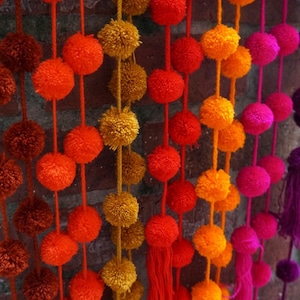 May include: A colorful garland of pom-poms in shades of orange, red, pink, and purple. The pom-poms are strung together on a string and hang from a brick wall.