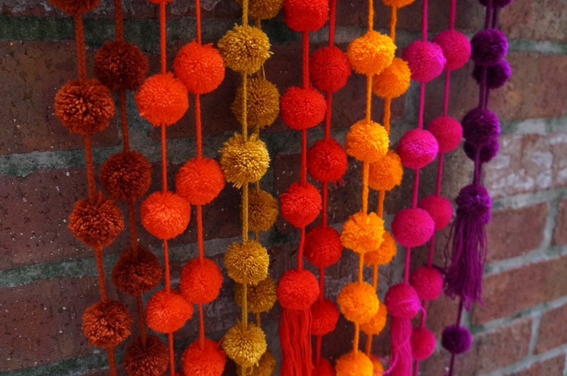 May include: A colorful garland of pom-poms in shades of orange, red, pink, and purple. The pom-poms are strung together on a string and hang from a brick wall.