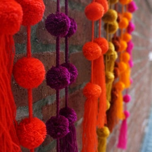 May include: A wall decorated with colorful pom-pom garlands. The garlands are made of fluffy, bright orange, pink, purple, and yellow pom-poms. Each pom-pom has a long tassel hanging from it.
