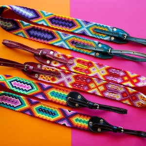 May include: Three colorful woven camera straps with leather accents. The straps feature geometric patterns in shades of red, orange, yellow, green, blue, purple, and white. The leather accents are brown and black.