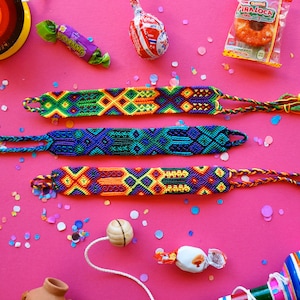 Könnte beinhalten: Drei bunte Freundschaftsarmbänder mit komplizierten Mustern in Blautönen, Grün, Gelb, Orange und Lila. Die Armbänder liegen auf einer rosa Oberfläche mit Konfetti und anderen festlichen Gegenständen.