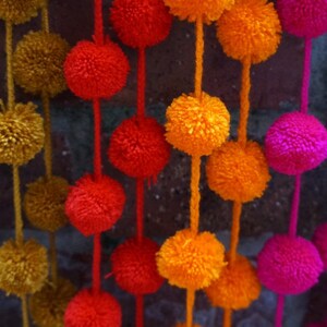 May include: A garland of colorful pom-poms in shades of orange, red, pink, and purple. The pom-poms are strung together on a white string and hang from a brick wall.