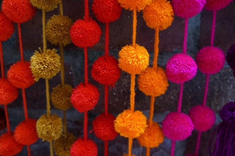 May include: A garland of colorful pom-poms in shades of orange, red, pink, and purple. The pom-poms are strung together on a white string and hang from a brick wall.