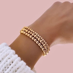 Könnte beinhalten: Drei goldene Perlenarmbänder auf einem Handgelenk. Die Armbänder bestehen aus kleinen, runden, glänzenden Perlen. Der Hintergrund ist rosa, und die Person trägt einen weißen Pullover.