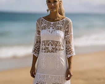 May include: White, sheer, lace cover-up dress with three-quarter sleeves. The dress features intricate floral lace patterns and a scoop neckline. The model is standing on a sandy beach with the ocean in the background.