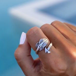 Könnte beinhalten: Ein silberfarbener Ring mit Feder- und Pfeil-Design. Der verstellbare Ring wird am Finger getragen und zeigt die detaillierte Textur der Feder und die Form des Pfeils.