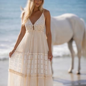May include: A cream-colored maxi dress with spaghetti straps and a V-neckline. The dress features lace detailing on the bodice and a tiered skirt with a geometric pattern. A white cowboy hat is worn. A white horse stands in the background.