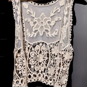 May include: A cream-colored crochet vest with a floral design. The vest is sleeveless and has a delicate, openwork pattern.