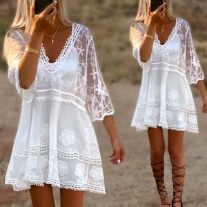 May include: White crochet mini dress with a V-neckline and 3/4 sleeves. The dress has a tiered design and a delicate lace trim.