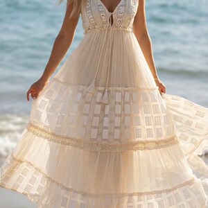 May include: A flowing, cream-colored maxi dress with spaghetti straps and a V-neckline. The dress features a tiered skirt with a geometric pattern and lace trim. A tassel detail hangs from the neckline. The dress is suitable for a beach or summer occasion.