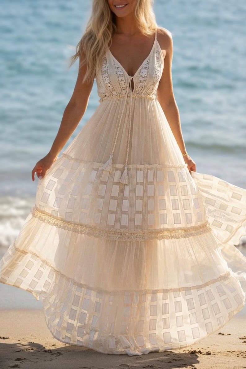 May include: A flowing, cream-colored maxi dress with spaghetti straps and a V-neckline. The dress features a tiered skirt with a geometric pattern and lace trim. A tassel detail hangs from the neckline. The dress is suitable for a beach or summer occasion.