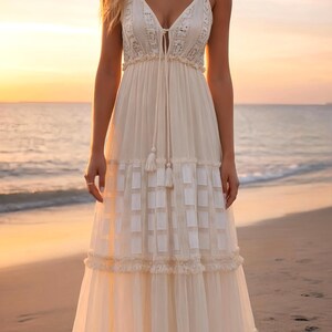 May include: A cream-colored maxi dress with spaghetti straps and a plunging neckline. The dress features a tiered skirt with a geometric pattern and delicate lace trim. The model is standing on a beach at sunset.