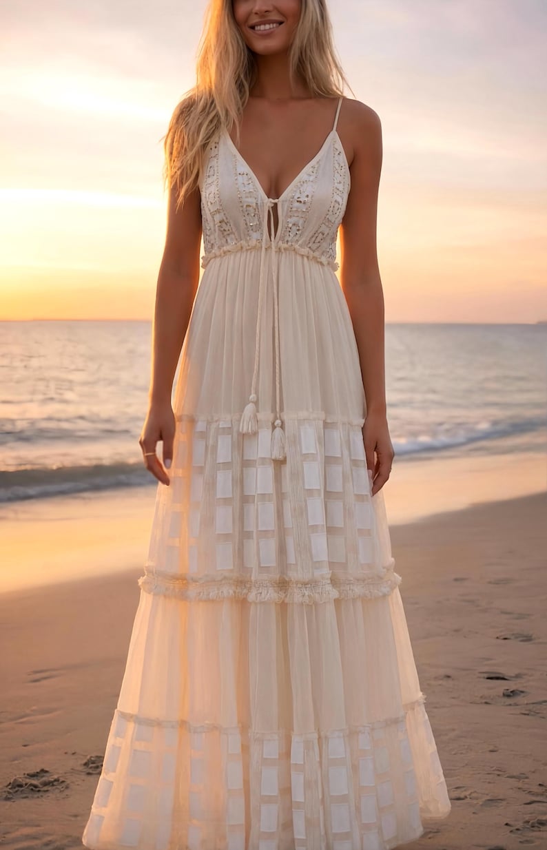 May include: A cream-colored maxi dress with spaghetti straps and a plunging neckline. The dress features a tiered skirt with a geometric pattern and delicate lace trim. The model is standing on a beach at sunset.