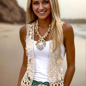 May include: A woman wearing a white tank top with a beige crochet vest, a green leather belt with a silver buckle, and a white lace skirt. She is wearing a necklace with white shell beads.