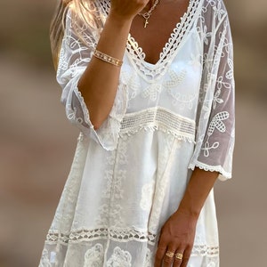 May include: White crochet mini dress with a V-neckline and 3/4 sleeves. The dress has a lace overlay and a scalloped hem.