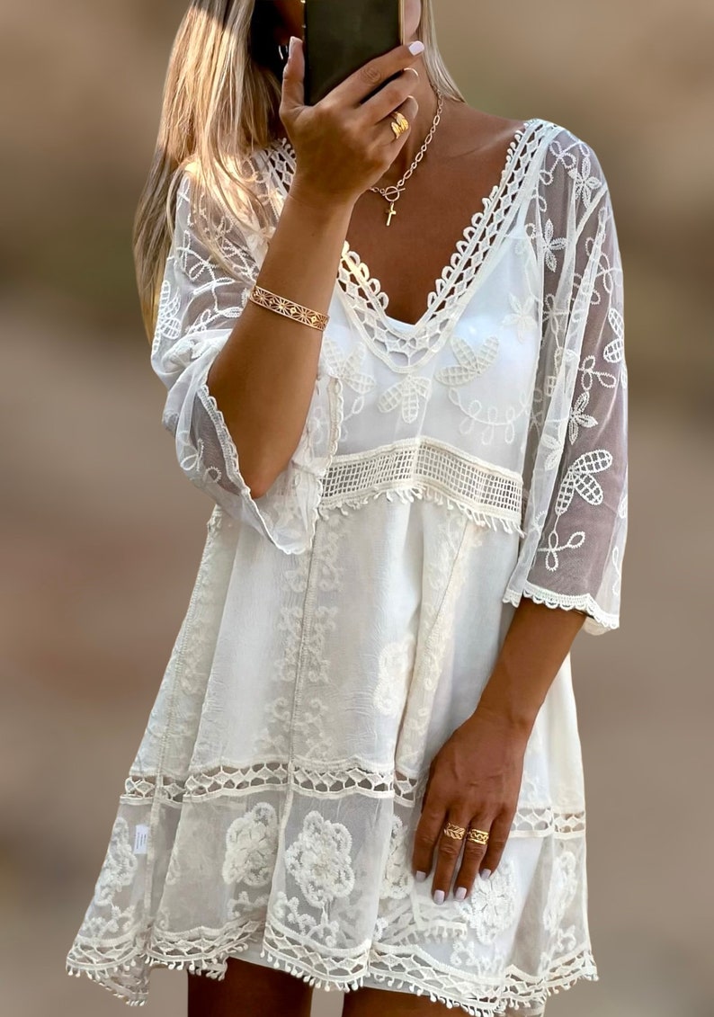 May include: White crochet mini dress with a V-neckline and 3/4 sleeves. The dress has a lace overlay and a scalloped hem.