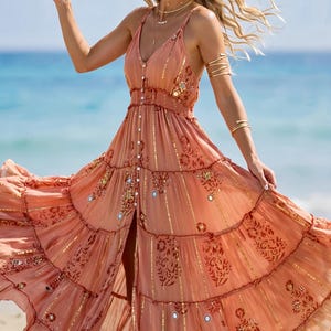 May include: A peach-colored maxi dress featuring a V-neckline, thin shoulder straps, and a button-down front. The fabric is adorned with metallic gold vertical stripes, floral patterns, and small circular mirror embellishments throughout.