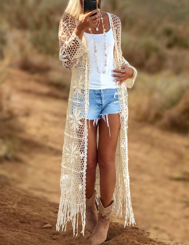 Puede incluir: Un c&aacute;rdigan largo de crochet de color crema con detalles de flecos. El c&aacute;rdigan est&aacute; abierto por delante y tiene un delicado patr&oacute;n floral. La modelo lleva el c&aacute;rdigan sobre un top blanco de tirantes y pantalones cortos de mezclilla.