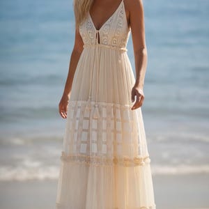 May include: A cream-colored maxi dress with a V-neckline and spaghetti straps. The dress features a tiered skirt with a grid pattern and lace trim. The model is standing on a beach with the ocean in the background.