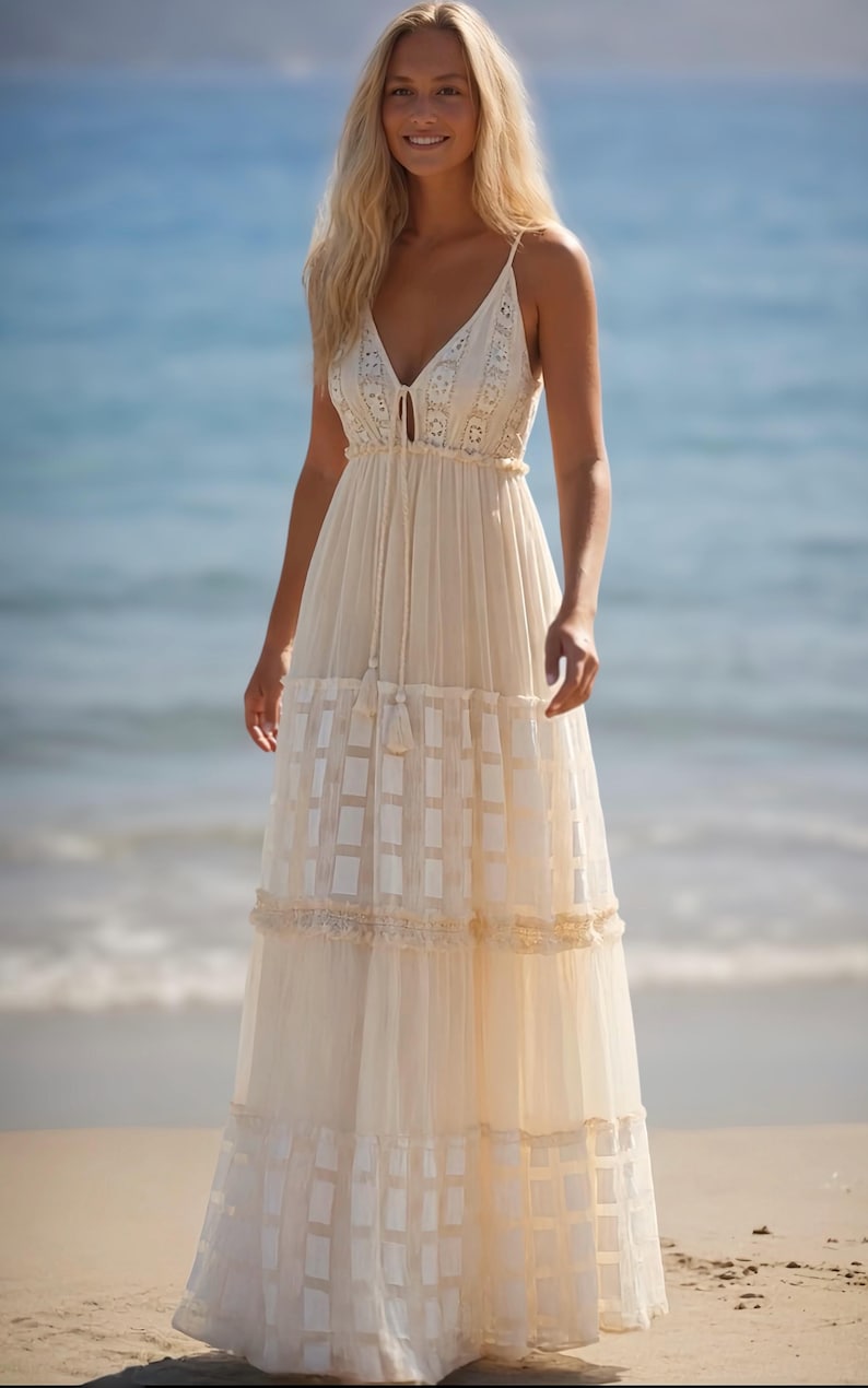May include: A cream-colored maxi dress with a V-neckline and spaghetti straps. The dress features a tiered skirt with a grid pattern and lace trim. The model is standing on a beach with the ocean in the background.