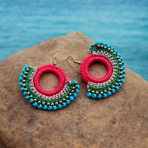 Può includere: Un paio di orecchini all'uncinetto in una combinazione di colori rosa, verde e beige. Gli orecchini sono decorati con perline turchesi e dorate.