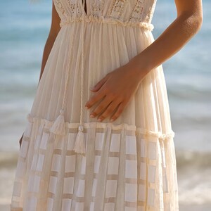 May include: Cream-colored dress with a V-neckline and intricate embroidered details. The dress features a tiered skirt with a geometric pattern and tassel accents. The lightweight fabric and flowing design create a bohemian aesthetic, perfect for warm weather.