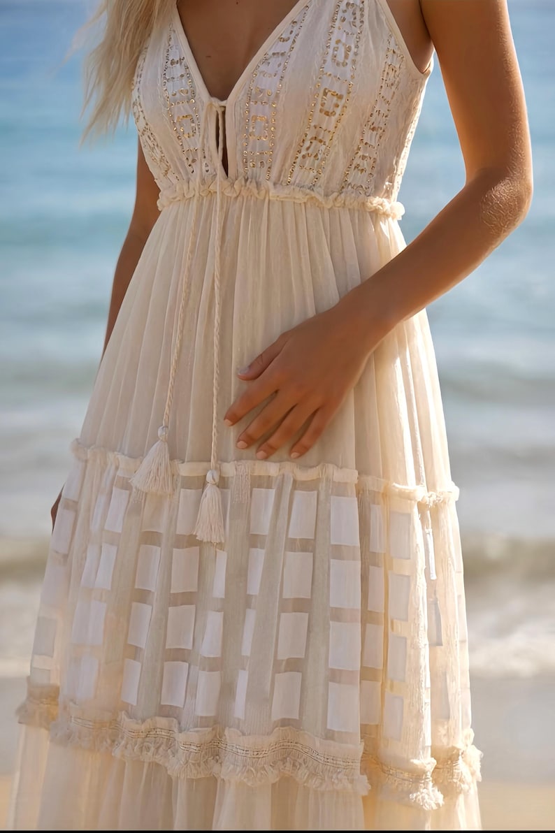 May include: Cream-colored dress with a V-neckline and intricate embroidered details. The dress features a tiered skirt with a geometric pattern and tassel accents. The lightweight fabric and flowing design create a bohemian aesthetic, perfect for warm weather.