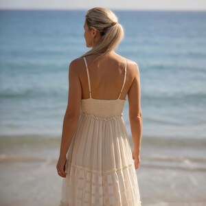 May include: A cream-colored sundress with spaghetti straps and a tiered skirt. The dress has a fitted bodice and a flowing, patterned skirt. The background features a beach and ocean.